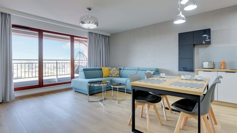 A spacious and open living room with a comfortable blue sectional sofa, a dining table, and large windows providing plenty of natural light.