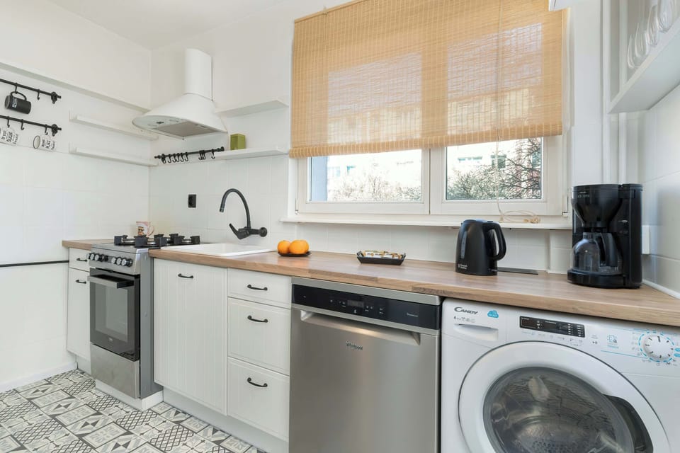 A kitchen with white cabinets and a wooden countertop. Various appliances stand on the countertop, and a window can be seen in the background. The style is modern with warm, natural accents.