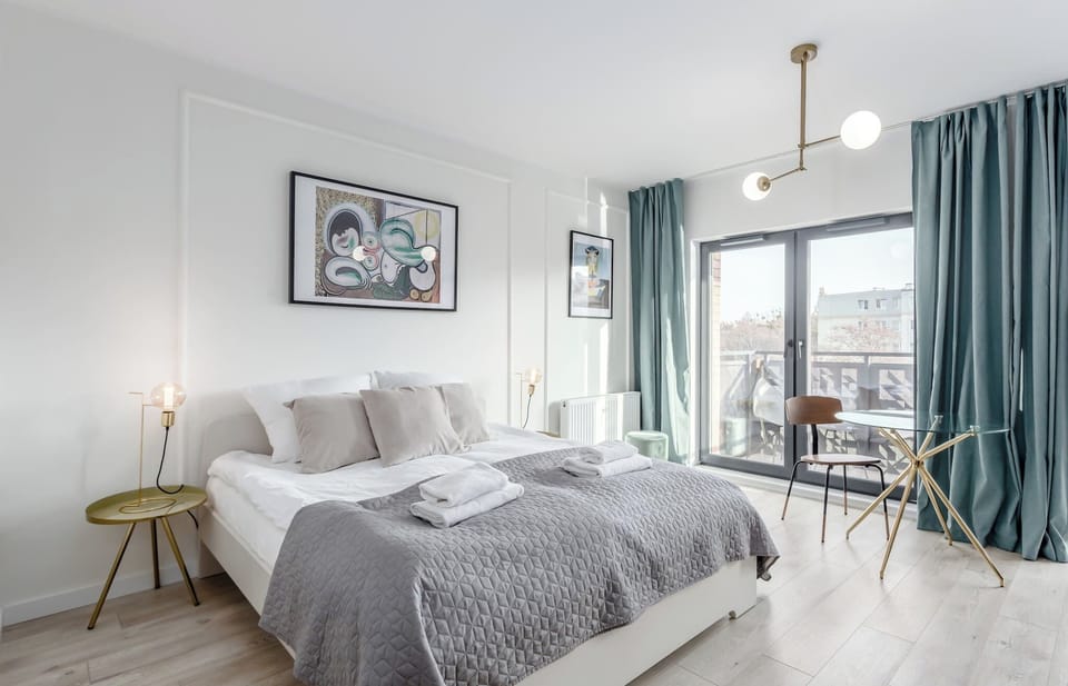 A spacious bedroom with a large bed, soft gray bedding, and a balcony view. Natural light streams in through wide windows with curtains.