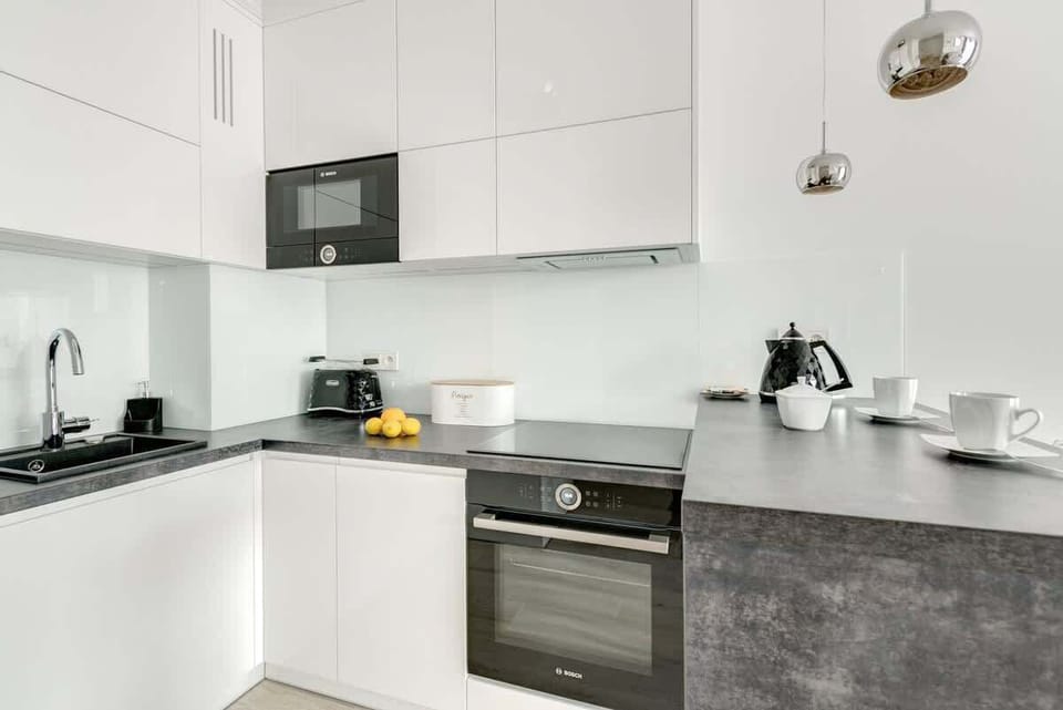 A modern, minimalist kitchen with sleek white cabinetry, a black sink, and built-in appliances, complemented by metallic pendant lights and a dark countertop.
