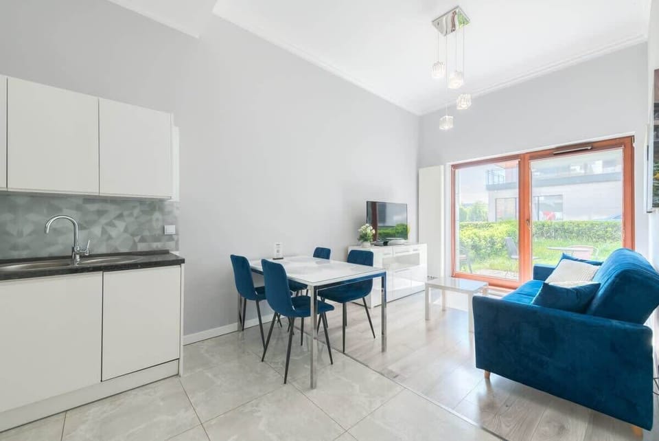 The modern apartment features a bright living area with a blue sofa, dining table, a sleek kitchen, and large windows opening to a garden view.