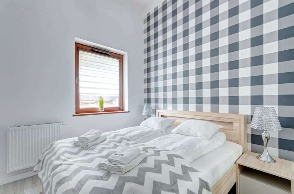 A bedroom with a wooden bed frame, a patterned gray and white duvet, two bedside tables, and a window with a potted plant on the windowsill.