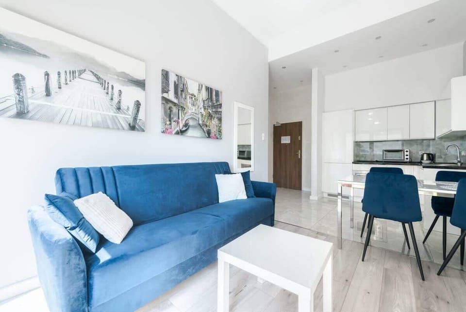 The living area showcases a vibrant blue sofa adorned with white cushions, modern wall art, and an open-plan kitchen in a bright, minimalist design.