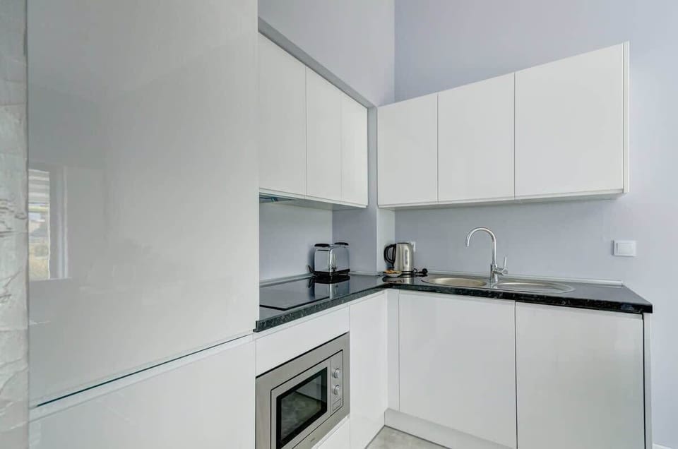 A modern white kitchen with a sink, a kettle, a toaster, a microwave, and storage cabinets, all set against a light-colored wall.