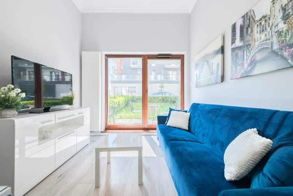 The cozy living room features a vibrant blue sofa with white cushions, a sleek white TV unit, and large glass doors opening to a garden view.