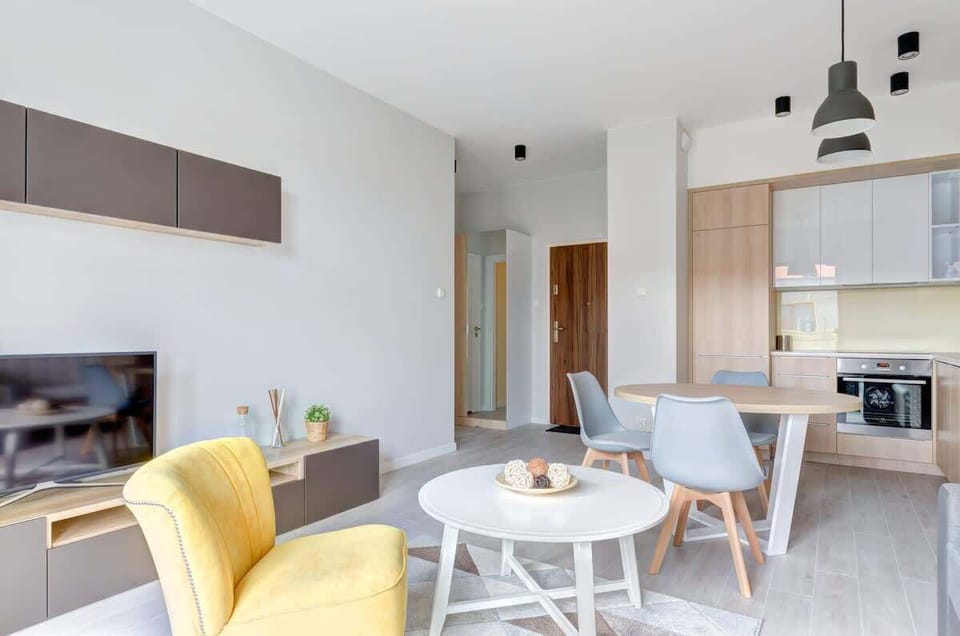 The cozy living space features a modern dining area, a yellow accent chair, a TV, and a kitchen with light wood finishes and sleek white cabinetry.