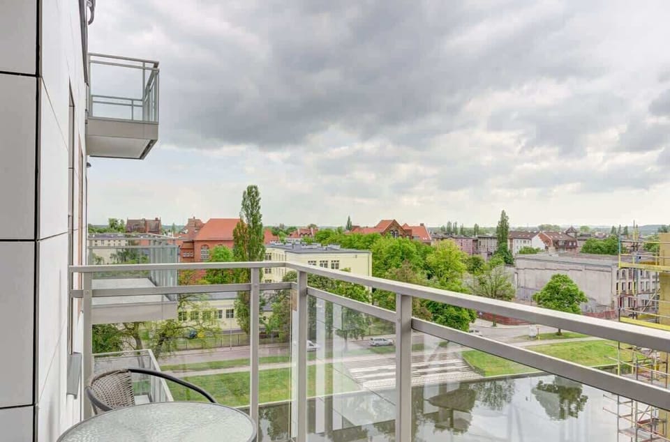 A view from a balcony with a table and chair, overlooking a cityscape filled with greenery, buildings, and partly cloudy skies.