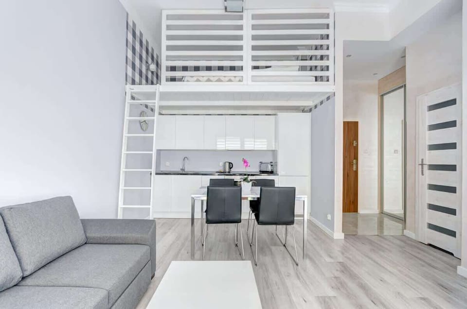 A modern, bright interior with a loft-style sleeping area accessible by a ladder, a minimalist white kitchen, a dining table with black chairs, and a gray sofa in the foreground.