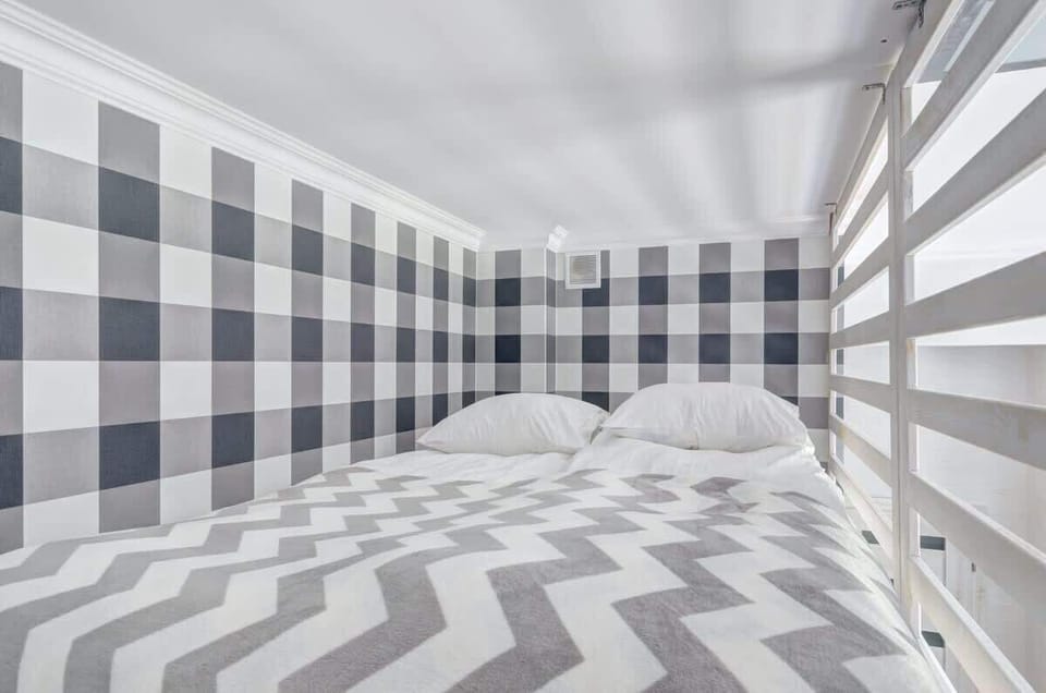 A cozy loft bed with white pillows, a patterned gray and white blanket, and walls decorated with a black and white checkered pattern.