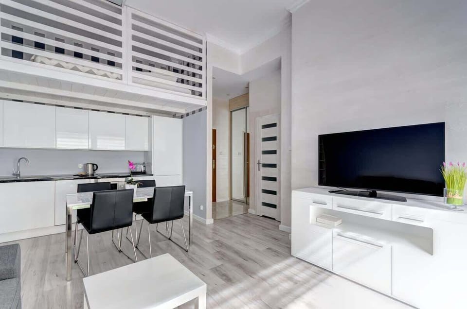 The interior features a bright, modern kitchen with sleek white cabinets, a dining area with black chairs, a loft-style sleeping space above, and a living room with a large TV and minimalist design.