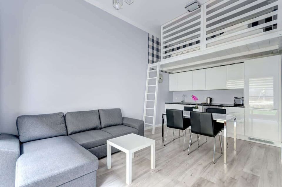 A modern studio apartment with a gray sectional sofa, a white coffee table, a sleek kitchen area, a dining table with black chairs, and a loft bed accessible via a white ladder.