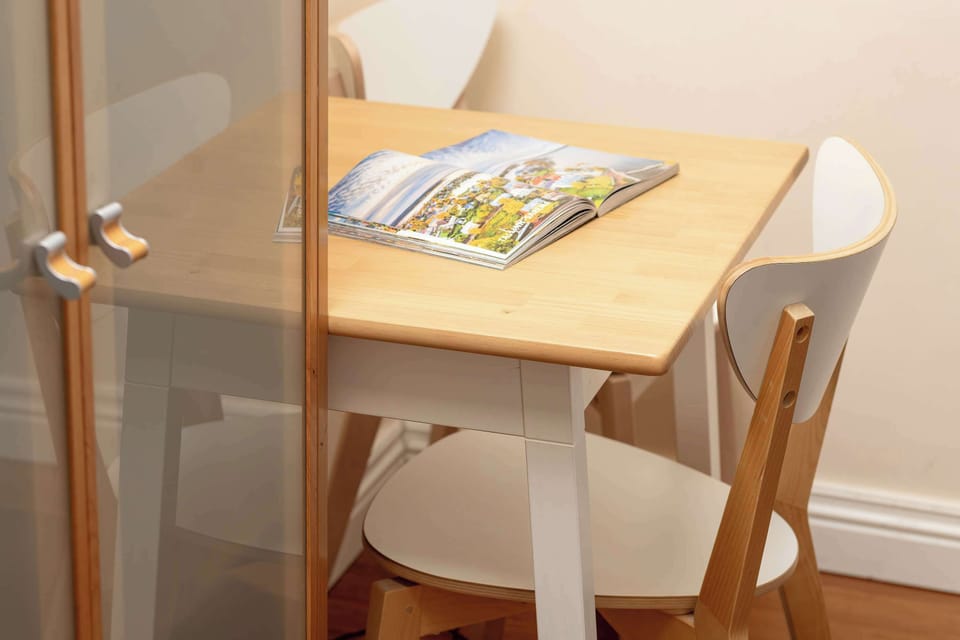 A small wooden table with a magazine and a chair placed next to a cream-colored wall inside the bedroom.

