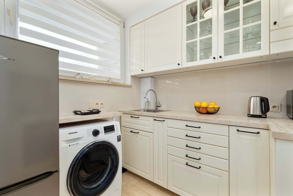 A full view of the kitchen showing a washing machine, fridge, oven, and upper cabinets beneath a wide window.


