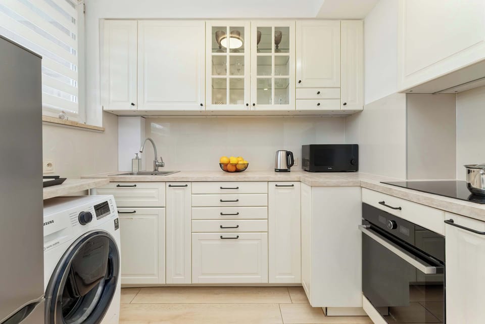 A modern kitchen with white cabinets, a washing machine, microwave, oven, and open shelving with dishes.

