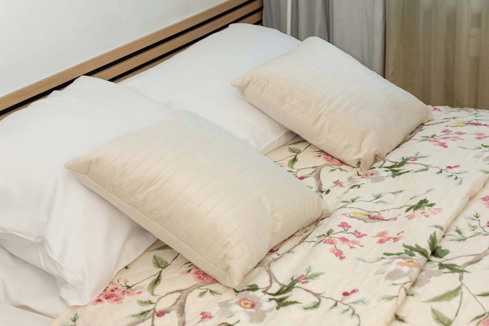 A detail shot of the double bed with decorative pillows and floral bedding, placed near a window with light curtains.

