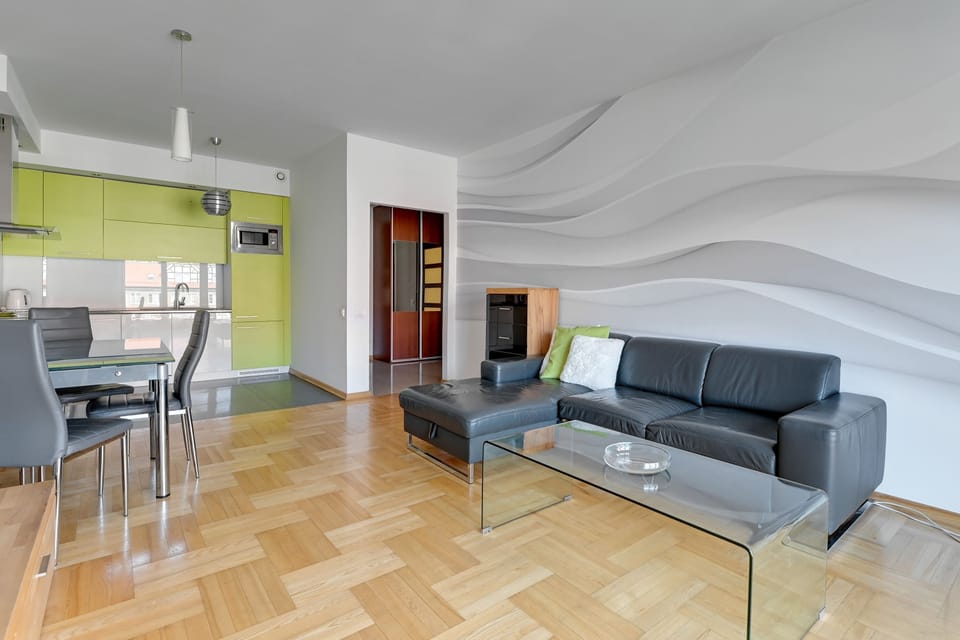 A spacious open-plan living area combining a living room and kitchen. The room is furnished with light gray seating and a dining table, enhanced by large windows that flood the space with natural light.
