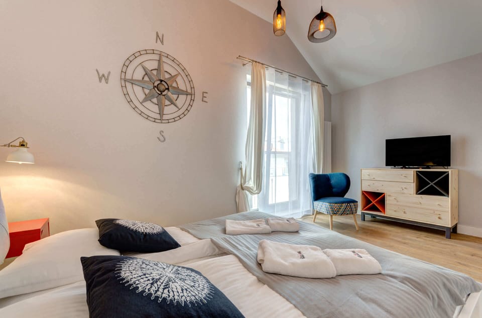 A cozy studio with a nautical theme, featuring a bed, sitting area, and large windows letting in natural light.