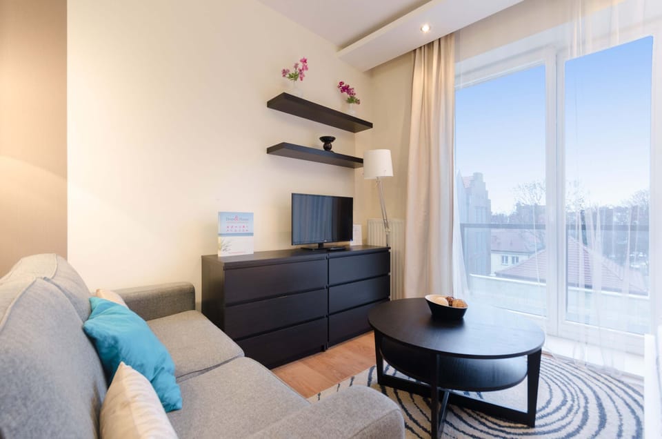 A cozy living room featuring a gray sofa, small black coffee table, and minimalist shelves with decorations. Large windows provide natural light.
