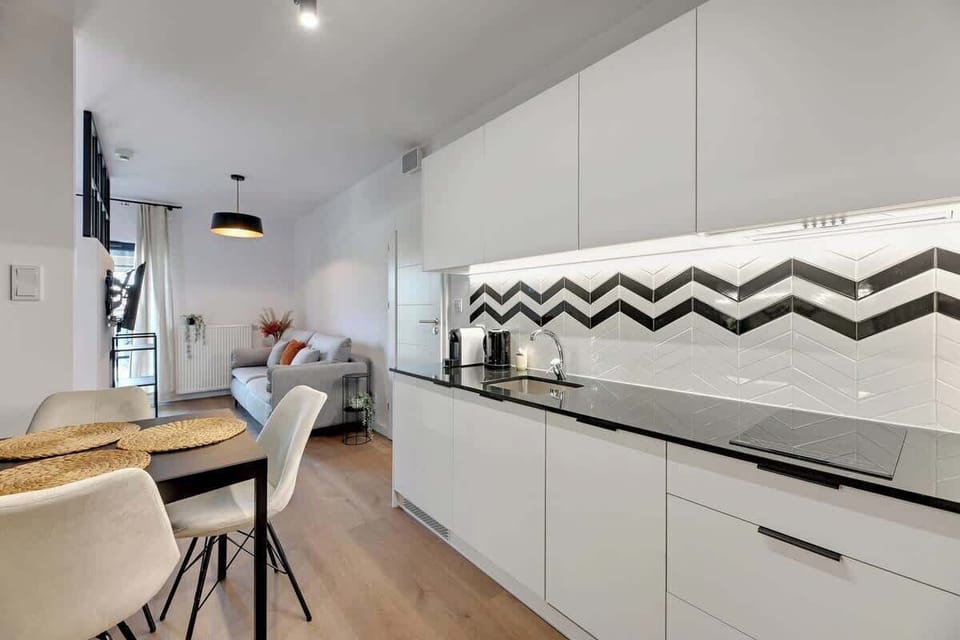 An elegant kitchenette with white furniture and a black and white pattern on the wall is complemented by a minimalist dining table. 
