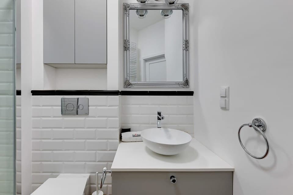 Modern fixtures, a refined silver-framed mirror, and an elegant minimalistic design, highlighted by smooth white tiles and contrasting black accents