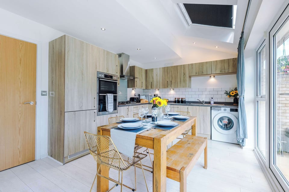 Bright, modern kitchen with full appliances, stylish wood cabinetry, and a spacious dining area. Enjoy meals at the rustic table with views of the garden through large French doors. Perfect for cooking, dining, and gathering.