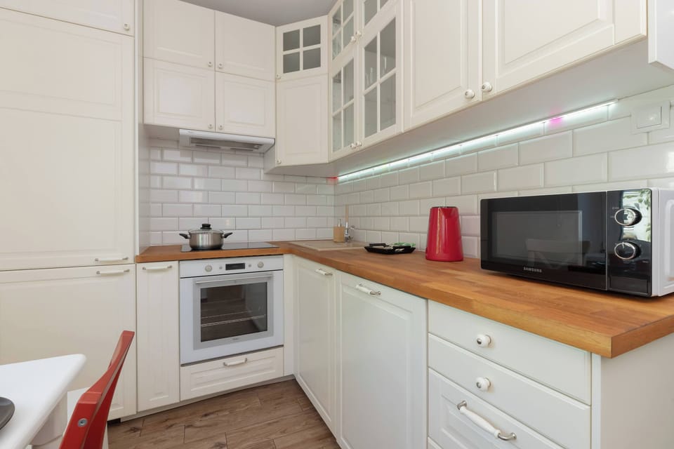 A wide shot of the kitchen showing white cabinets, microwave, red kettle, and a tiled backsplash.

