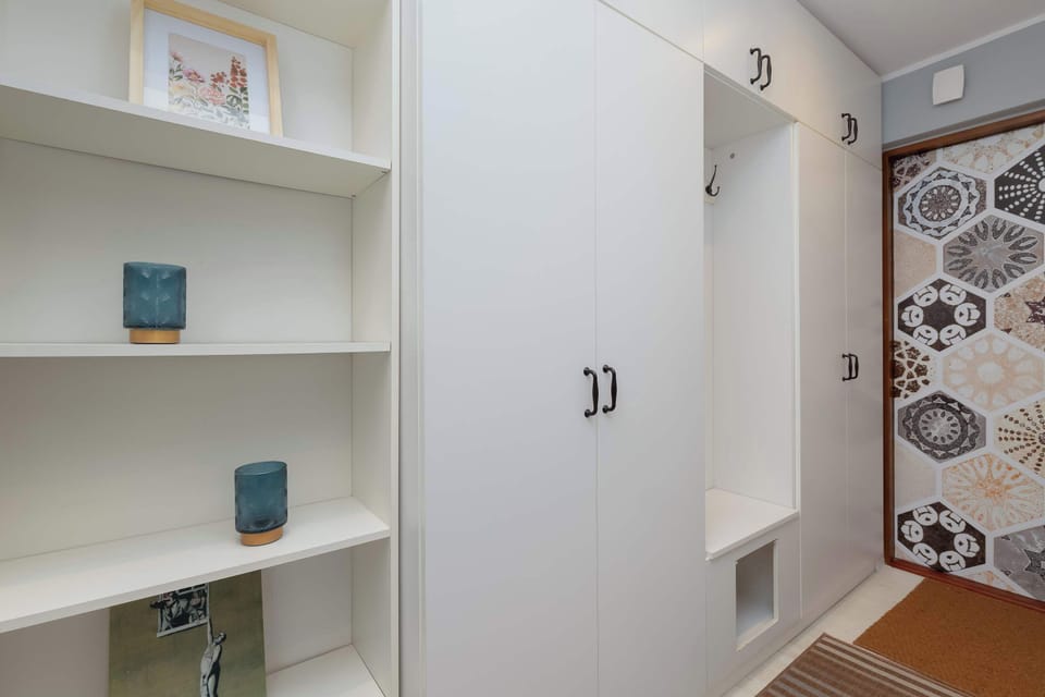 A bright entryway with open shelving, white closets, and a glimpse of the patterned bathroom beyond.