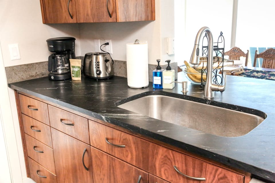 This view highlights the stunning contrast between the sleek, dark stone countertop and the richly-grained, warm wood cabinetry. The undermount stainless steel sink and modern gooseneck faucet are complemented by essential amenities like the coffee station