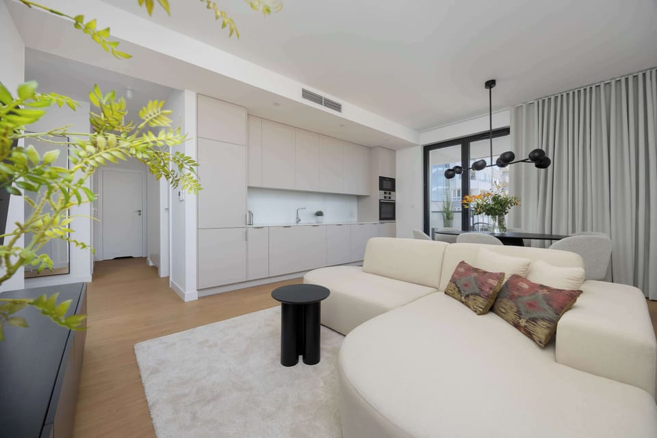 A modern living room with a soft beige sofa, minimalist decor, and a view into the kitchen area with bright natural light.