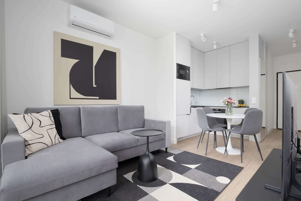 A bright living room featuring a sectional sofa, small dining table, and white interior palette with minimalist decor.