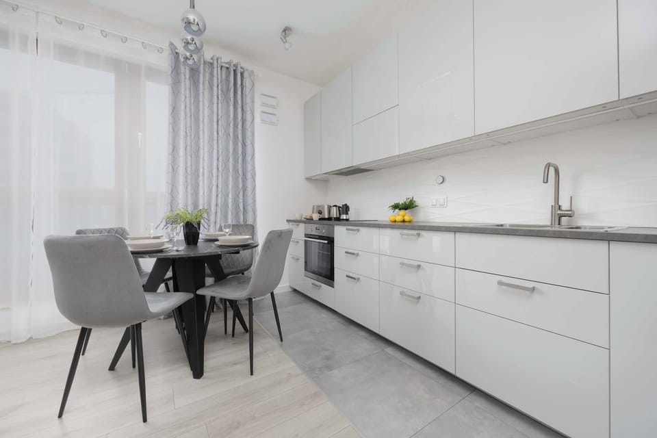 A well-lit open kitchen area with white cabinetry, a gray backsplash, and a dining table with gray chairs. The room is modern and practical.
