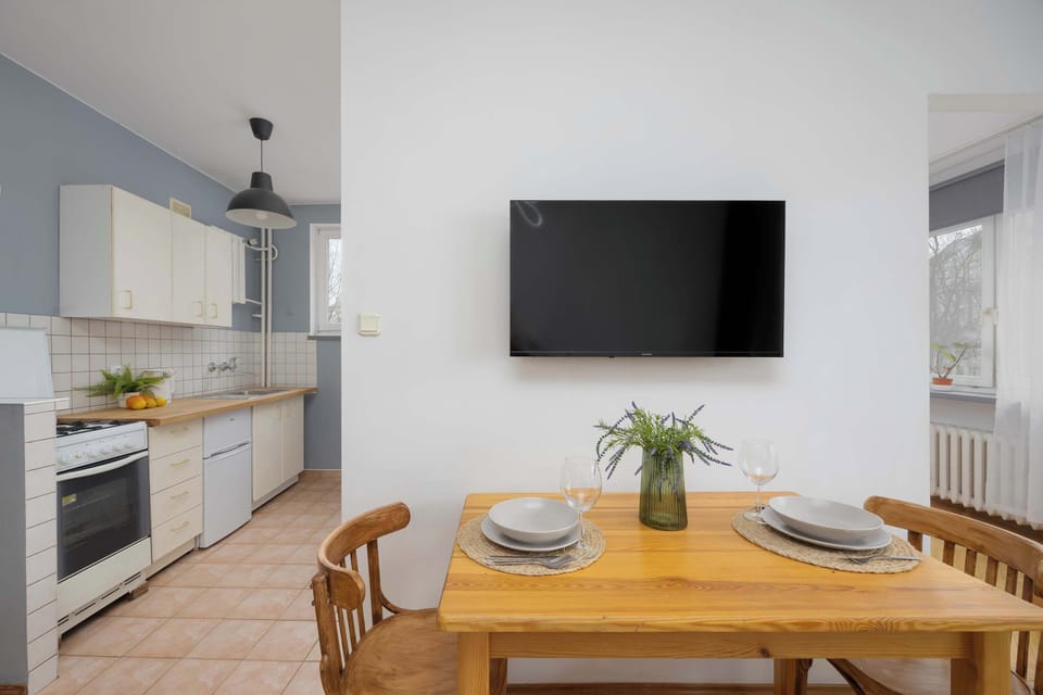 A bright living room with a wooden dining table, and a TV mounted on the wall. The open space connects to the kitchenette.
