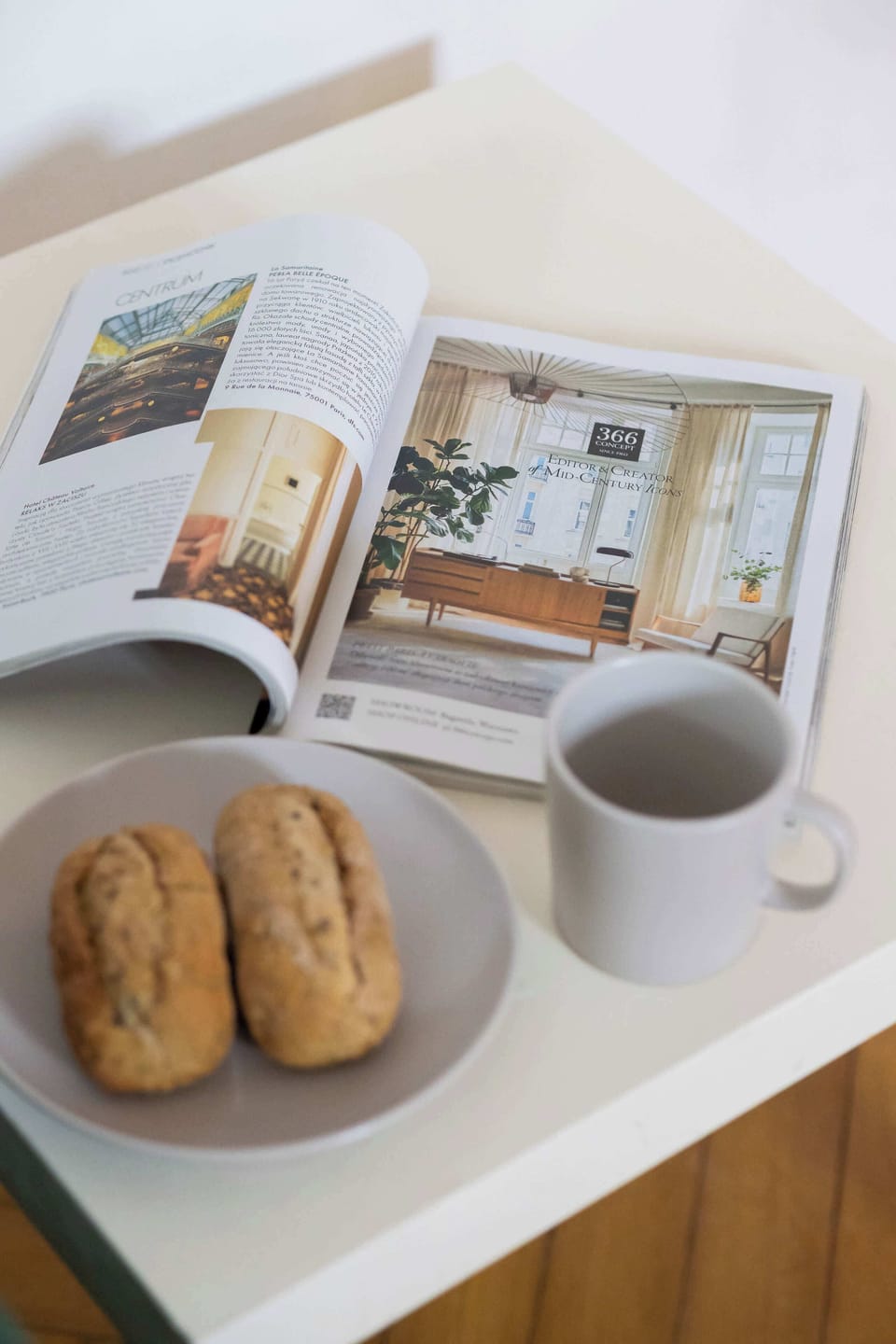 A breakfast setup on a white table, featuring a newspaper, a cup of coffee, and two small bread rolls.
