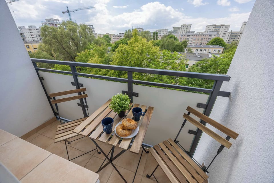 A narrow balcony with tile flooring, folding chairs, a small table, and fresh drinks set against a green urban backdrop.