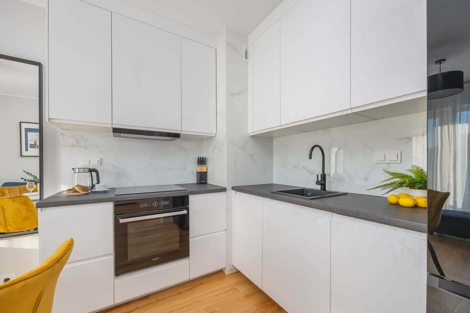 A clean, white kitchenette with minimalistic cabinetry, wooden flooring, and a few decorative touches.