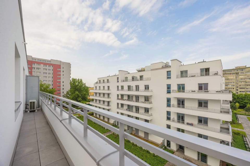 A long balcony view reveals modern buildings, lush greenery, and city surroundings under a bright sky.

