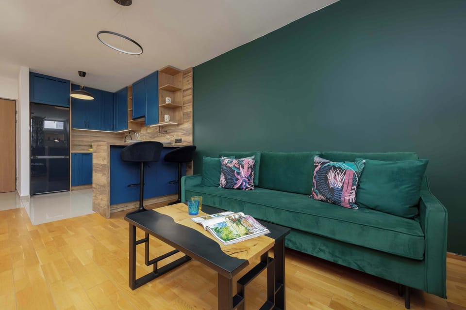 A living room with a view of the open kitchen area. The room has a minimalist style with neutral wood tones.