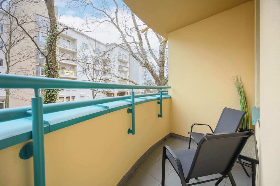 A balcony with a green railing, overlooking the outdoor environment with chairs visible.