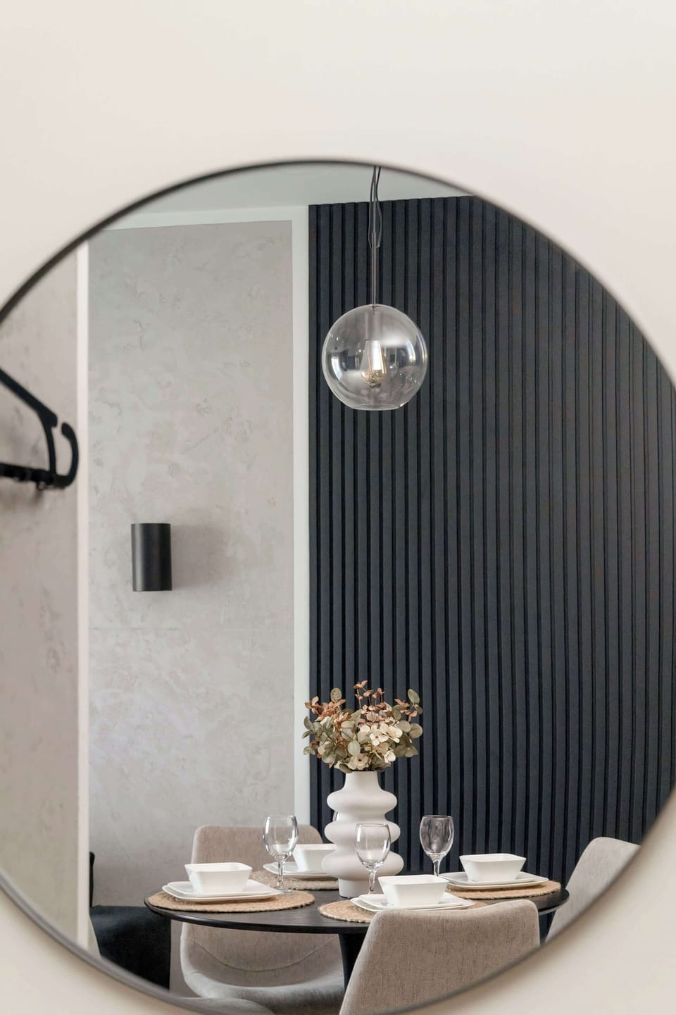 A close-up of a round wall mirror above a dining table with a simple floral arrangement and monochrome tones.

