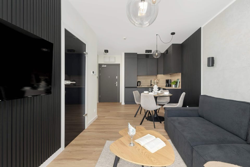 A long shot of a living space with a grey couch, wooden flooring, and black-accented kitchen in the background.