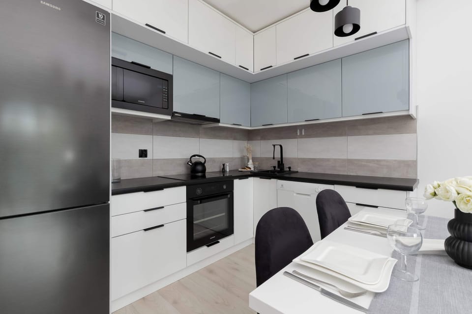 A modern kitchenette, with white cabinets and a dark countertop, with a microwave, kettle and oven.