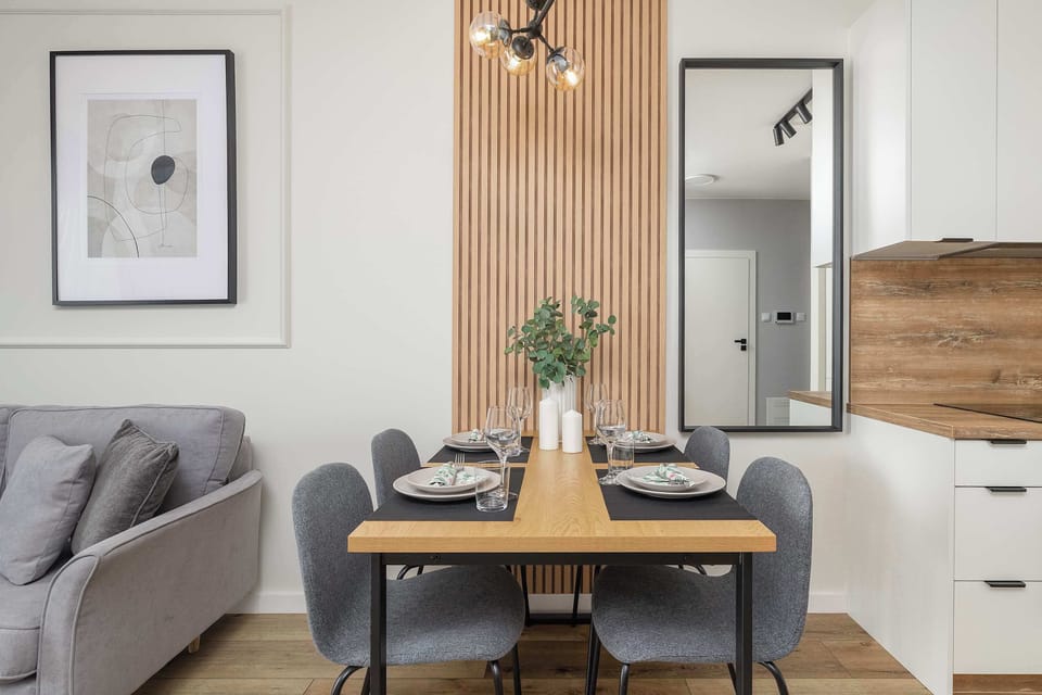 A wider shot of the dining and kitchen area with white cabinetry, a wooden backsplash, and a sleek black dining table. Large windows and sliding doors bring in ample natural light.

