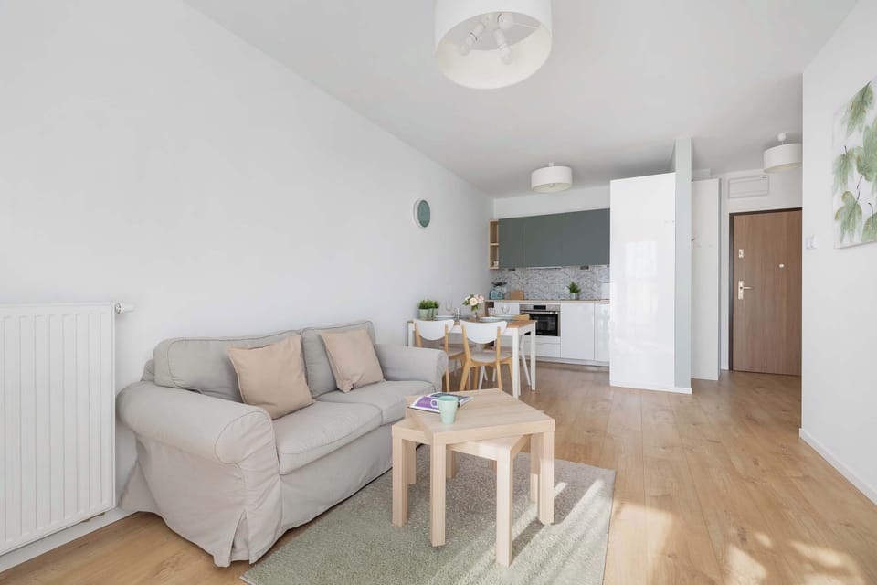 Spacious living room with a large gray sofa, coffee table, and mounted TV. Light curtains bring in natural light, creating a cozy atmosphere.