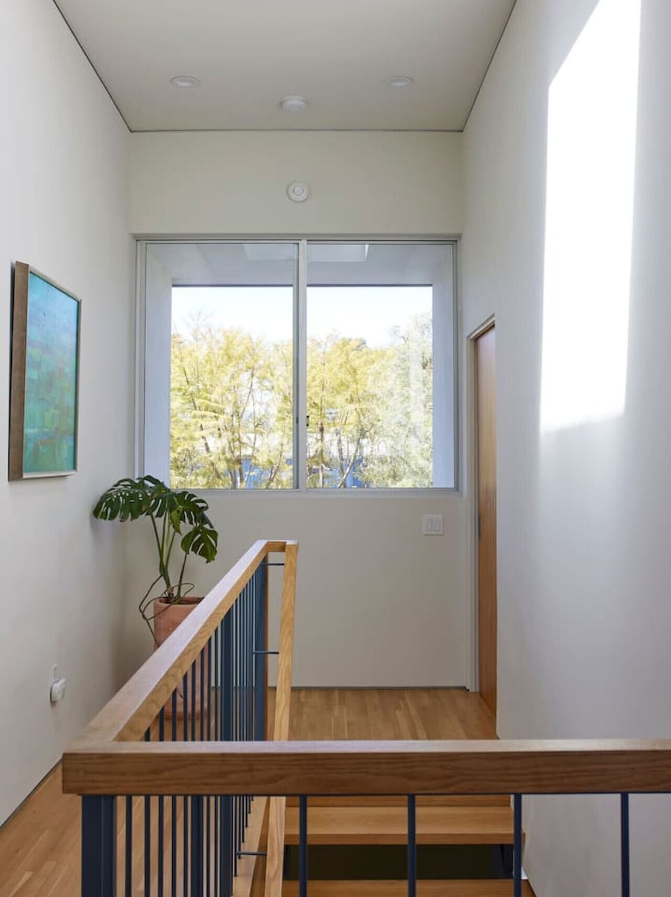 A clean, airy landing with natural wood accents, sleek metal railing, and a bold teal artwork. Skylight above brings in soft daylight for a fresh, open feel.