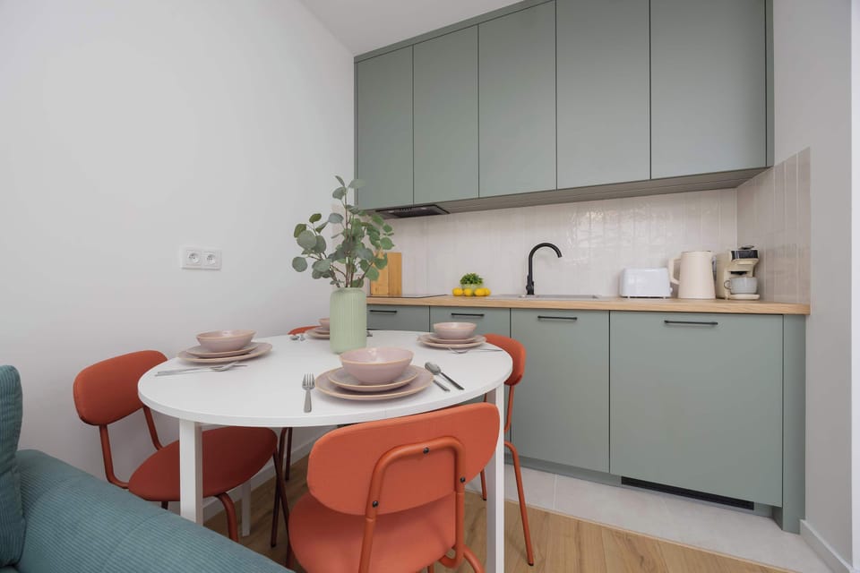 A compact dining area with a round white table and four coral-colored chairs, adjacent to a light green kitchenette.

