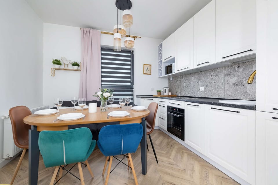 Table with colourful chairs, tableware and flowers. A white kitchen with dark worktop visible in the background.