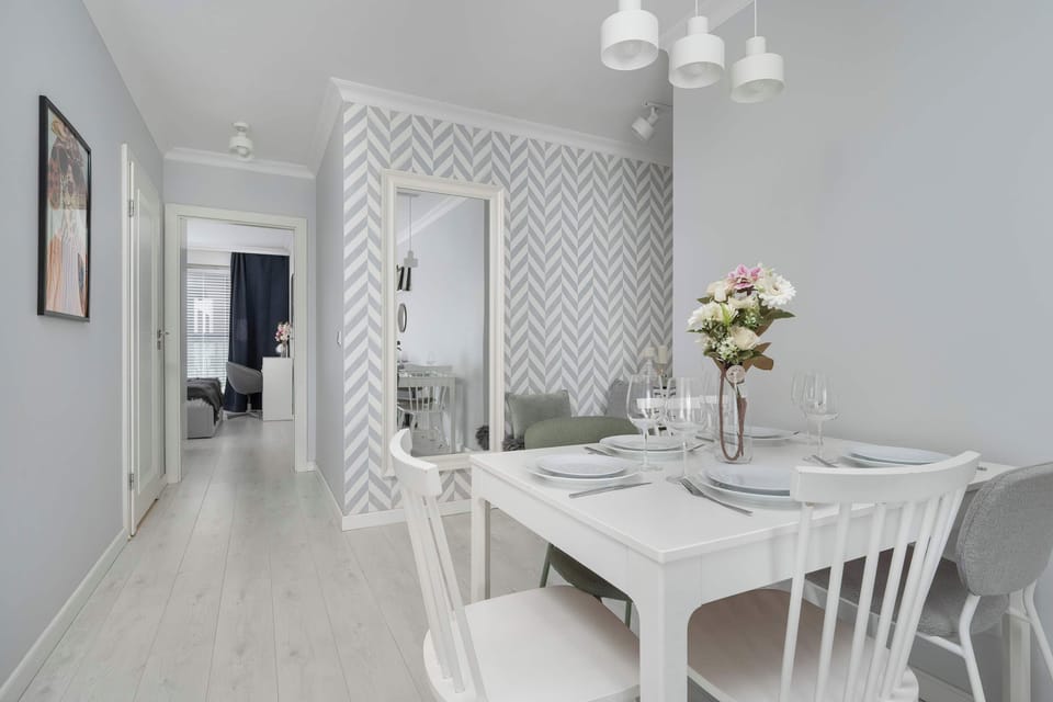 A dining area featuring a round white table with matching chairs, light wooden floors, and a patterned wall with a flower arrangement.