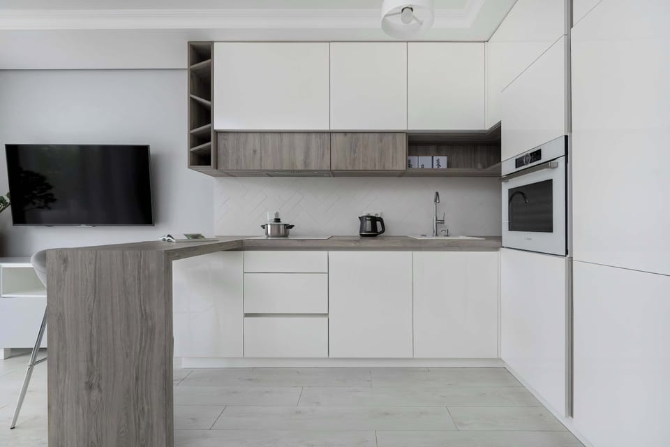 A sleek kitchen with white cabinets, built-in appliances, and a mounted TV on the wall across from a bar table.