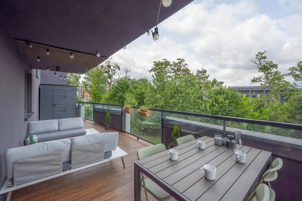A covered balcony with a modern outdoor sofa, cushions, and a glass railing overlooking green trees.