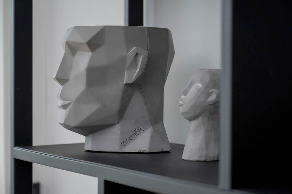 A modern sculpture of a geometric human head placed next to a classic bust on a black shelf.
